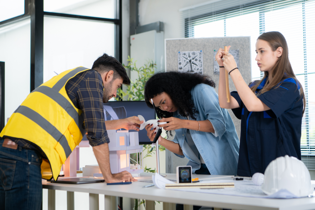 Group of engineers working on a model, measuring, and discussing details in a bright workspace with design tools and equipment.