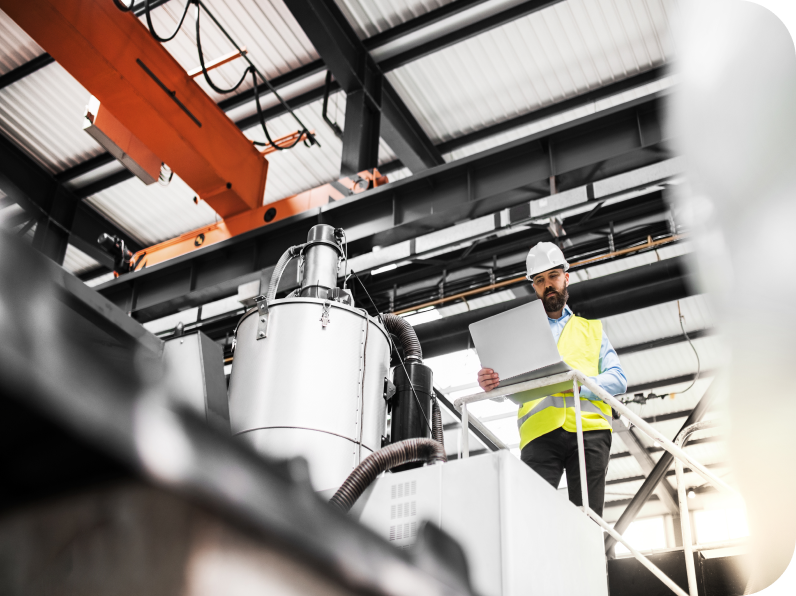 Engineer using a laptop while monitoring large machinery in an industrial facility, wearing a hard hat and safety vest.