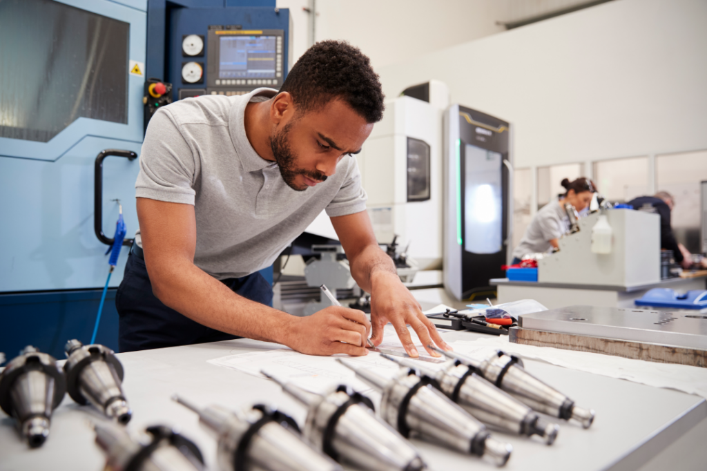 Engineer focused on technical drawings, with precision components and CNC machines in the background in a high-tech manufacturing facility.