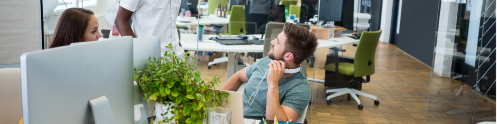 Two professionals having a discussion at a desk in a modern office with open desks, plants, and bright natural lighting.