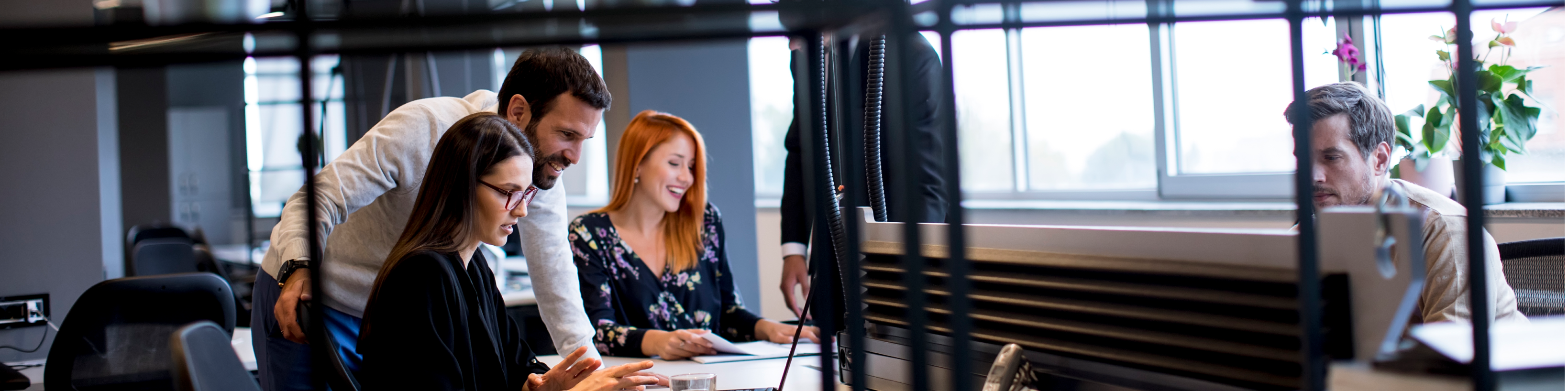 Group of professionals collaborating at a desk in a bright office with large windows, discussing ideas and working together.